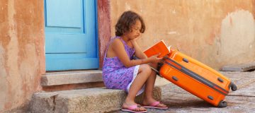 Una niña con uno de sus libros para el verano y una maleta sentada en un portal de un pueblecito.