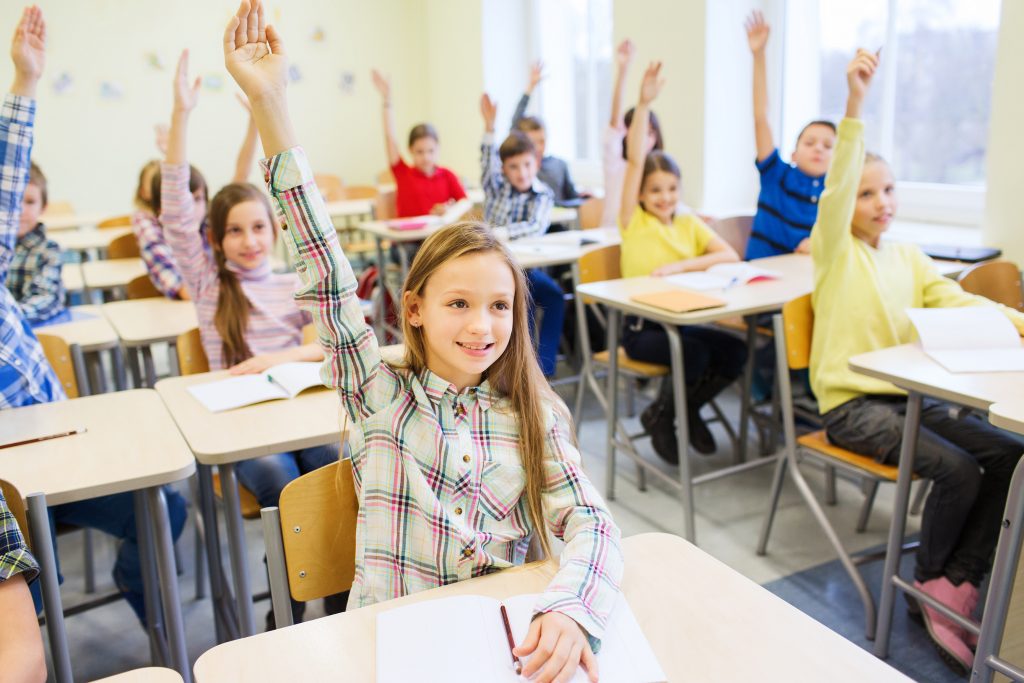 Grupo de niños en sus pupitres de clase con la mano alzada para ilustrar la actividad de hacer filosofía